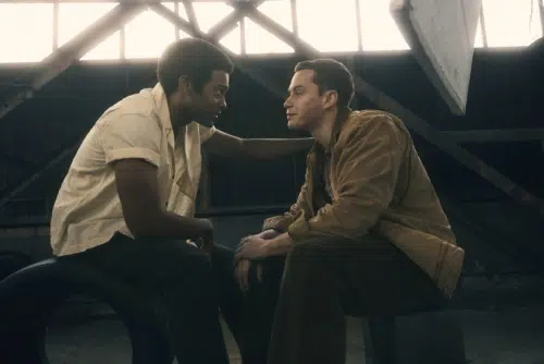 Dallas, played by Joshua Boone, and Darrel, played by Brent Comer, are sitting and facing each other in an industrial setting with large windows and metal beams in the background. Dallas, dressed in a short-sleeved shirt, has is left arm extended and his hand is on Darrel's shoulder, who is wearing a brown jacket. Both are engaged in a serious conversation, with intent expressions on their faces.