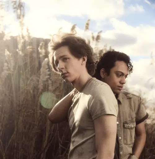 Ponyboy, played by Brody Grant, and Johnny, played by Sky Lakota-Lynch, stand in front of tall, dry grass with the sky and clouds in the background. Ponyboy runs a hand through his tousled hair, wearing a light-colored t-shirt, while Johnny stands slightly behind him, wearing a buttoned-down shirt with rolled-up sleeves. The image has a warm, soft focus with a light lens flare, creating a dreamy, contemplative atmosphere.