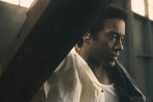 Dallas, played by Joshua Boone, with short, curly hair, looking contemplatively to the side in a dimly lit industrial setting. Dallas is wearing a white shirt with the collar slightly turned up and a dark jacket draped over one shoulder.