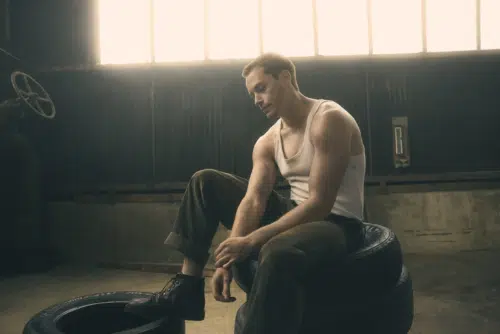 Darrel, played by Brent Comer, is wearing a white tank top and rolled-up pants. He is sitting on a stack of tires in a dimly lit industrial setting, next to a large wheel valve.