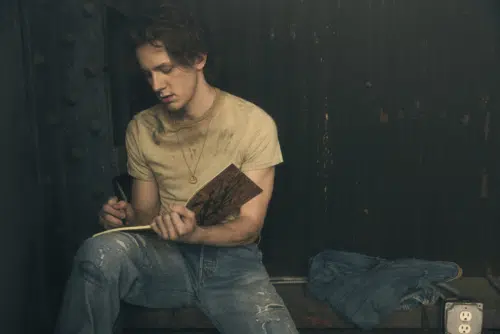 Ponyboy, played by Brody Grant, sits in a dimly lit industrial setting, wearing a dirty yellow t-shirt and distressed jeans. He holds a notebook and pen, appearing deep in thought as he writes. A denim jacket is draped next to him on the metal surface.