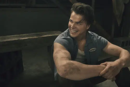 Sodapop, played by Jason Schmidt, smiles while sitting with his hands clasped around his knees. He is wearing a blue short-sleeved shirt with a name patch reading "Sodapop," a white undershirt, and a necklace. Their arms are dirtied, suggesting they have been working.