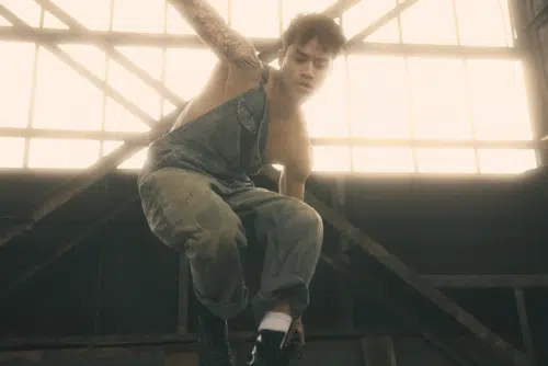 Two-Bit, played by Daryl Tofa, wearing distressed denim overalls and black boots, is captured in motion against a backdrop of large industrial windows. The lighting creates a warm glow, highlighting their muscular build and the geometric tattoo on their arm. The individual appears to be engaged in an action or dance pose inside an industrial setting.