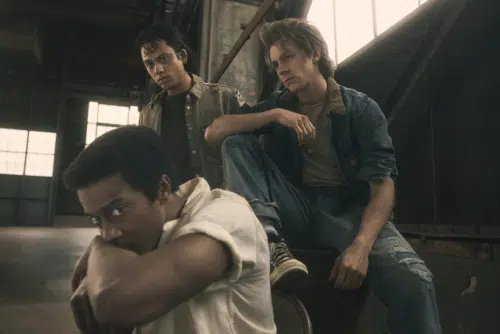 Dallas, Johnny, and Ponyboy pose in a gritty, industrial setting with natural light streaming in. Dallas, played by Joshua Boone, in the foreground rests his chin on his arm, looking serious. Johnny, played by Sky Lakota-Lynch, wears a military-style jacket, appearing contemplative. Ponyboy, played by Brody Grant, seated in a denim jacket and ripped jeans, looks thoughtful.