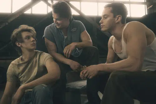 Ponyboy, Sodapop, and Darrel are sitting together in a dimly lit space with a large window in the background. Ponyboy, played by Brody Grant, has tousled hair and is wearing a light-colored t-shirt, looking to his right. Sodapop, played by Jason Schmidt, has dark hair, a blue button-up shirt with a name tag that reads 'Sodapop,' and is smiling while looking at Darrel. Darrel, played by Brent Comer, is wearing a white tank top and is leaning forward, looking at Sodapop. The setting appears industrial, with metal beams and a muted, vintage atmosphere.