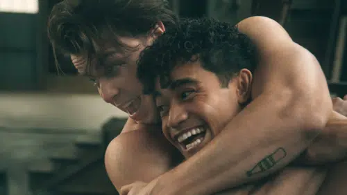 Sodapop and Two-Bit, engage in playful roughhousing. Sodapop, played by Jason Schmidt, with tousled hair, has a wide, animated expression. Two-Bit, played by Daryl Tofa, with curly hair, smiles broadly as he wraps his arm around his friend's neck.