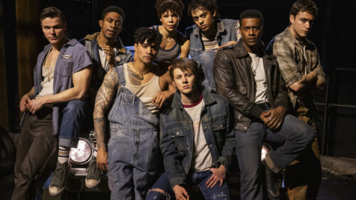 A group of eight young people, dressed in casual, rugged clothing, are posing together in front of a car in a dimly lit setting. Some are wearing denim jackets and overalls, while others sport leather jackets and plaid shirts. They all have serious or neutral expressions, embodying a tough, united group. The background is dark, highlighting the characters and their attire.