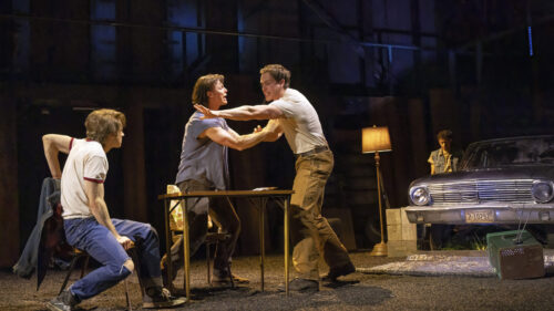 Ponyboy, Sodapop, and Darrel are engaged in a heated argument in a dimly lit, rustic setting. Darrel, played by Brent Comer, is being restrained by Sodapop, played by Jason Schmidt, who is standing and pushing him back. Ponyboy, played by Brody Grant, is seated and looks on intently. In the background, a vintage car and a floor lamp are visible, adding to the gritty, mid-20th-century atmosphere. Johnny stands near the car, looking down while the scene unfolds.