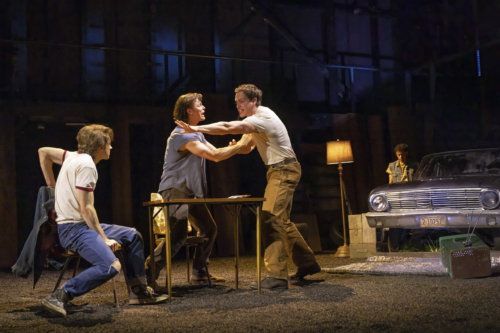 Ponyboy, Sodapop, and Darrel are engaged in a heated argument in a dimly lit, rustic setting. Darrel, played by Brent Comer, is being restrained by Sodapop, played by Jason Schmidt, who is standing and pushing him back. Ponyboy, played by Brody Grant, is seated and looks on intently. In the background, a vintage car and a floor lamp are visible, adding to the gritty, mid-20th-century atmosphere. Johnny stands near the car, looking down while the scene unfolds.