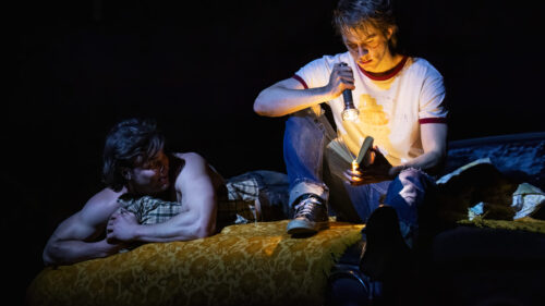 Ponyboy and Sodapop are in a dark setting, with Sodapop lying on a bed and Ponyboy sitting beside him, holding a flashlight and reading a book. Sodapop, played by Jason Schmidt, is shirtless and resting his head on a pillow and watching Ponyboy intently. Ponyboy, played by Brody Grant, is wearing a white t-shirt with red trim and jeans, focusing on the book illuminated by the flashlight.