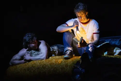 Ponyboy and Sodapop are in a dark setting, with Sodapop lying on a bed and Ponyboy sitting beside him, holding a flashlight and reading a book. Sodapop, played by Jason Schmidt, is shirtless and resting his head on a pillow and watching Ponyboy intently. Ponyboy, played by Brody Grant, is wearing a white t-shirt with red trim and jeans, focusing on the book illuminated by the flashlight.