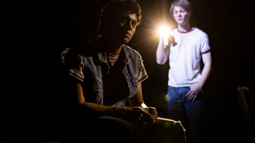 In a dark setting, Johnny and Ponyboy are highlighted by a bright light. Johnny, played by Sky Lakota-Lynch, is sitting, wearing a denim vest over a striped shirt, with his face partially obscured by shadows. Ponyboy, played by Brody Grant, wearing a white t-shirt with red trim and jeans, is standing and holding a flashlight, which casts a strong beam of light, illuminating his face and creating a dramatic atmosphere.
