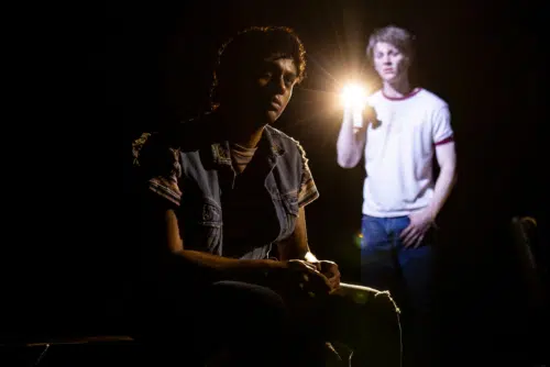 In a dark setting, Johnny and Ponyboy are highlighted by a bright light. Johnny, played by Sky Lakota-Lynch, is sitting, wearing a denim vest over a striped shirt, with his face partially obscured by shadows. Ponyboy, played by Brody Grant, wearing a white t-shirt with red trim and jeans, is standing and holding a flashlight, which casts a strong beam of light, illuminating his face and creating a dramatic atmosphere.
