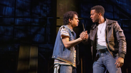 Johnny and Dallas are engaged in a tense confrontation in a dark setting. Johnny, played by Sky Lakota-Lynch, wearing a denim vest over a striped shirt, holds a knife, while Dallas, played by Joshua Boone, dressed in a leather jacket and white t-shirt, grips his wrist firmly. Their expressions are intense and serious, reflecting the gravity of the moment. The background is dimly lit, adding to the dramatic and suspenseful atmosphere.
