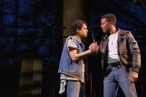 Johnny and Dallas are engaged in a tense confrontation in a dark setting. Johnny, played by Sky Lakota-Lynch, wearing a denim vest over a striped shirt, holds a knife, while Dallas, played by Joshua Boone, dressed in a leather jacket and white t-shirt, grips his wrist firmly. Their expressions are intense and serious, reflecting the gravity of the moment. The background is dimly lit, adding to the dramatic and suspenseful atmosphere.