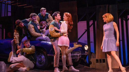 A group of young people are gathered around a blue car in a nighttime setting. Five young men, wearing matching letterman jackets, are either seated on or around the car, with one holding a football. Two young women are also present. Cherry, played by Emma Pittman, with long red hair and dressed in a pink skirt and white top is embracing one of the young men in a letterman jacket, while the other woman, dressed in a light blue dress, stands nearby, looking at them.