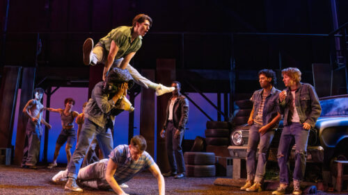 A lively scene unfolds with several young people engaged in an energetic dance or play fight. One man is leaping over another person covering their head with his letterman jacket. Other young men in casual, rugged clothing are either watching, dancing, or interacting in the background. The setting includes a vintage car and a stack of tires, with the backdrop featuring a dark, industrial structure and vibrant stage lighting.