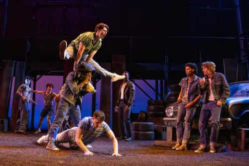 A lively scene unfolds with several young people engaged in an energetic dance or play fight. One man is leaping over another person covering their head with his letterman jacket. Other young men in casual, rugged clothing are either watching, dancing, or interacting in the background. The setting includes a vintage car and a stack of tires, with the backdrop featuring a dark, industrial structure and vibrant stage lighting.