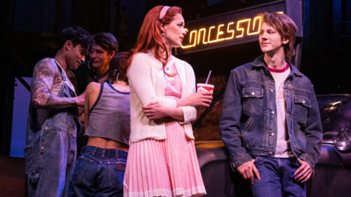 Cherry, played by Emma Pittman, dressed in a pink checkered dress and white cardigan, stands holding a drink cup with a straw. She is looking at Ponyboy, played by Brody Grant, in a denim jacket and jeans, who is leaning against a car and smiling at her. Behind them, three other young people are engaged in a conversation.