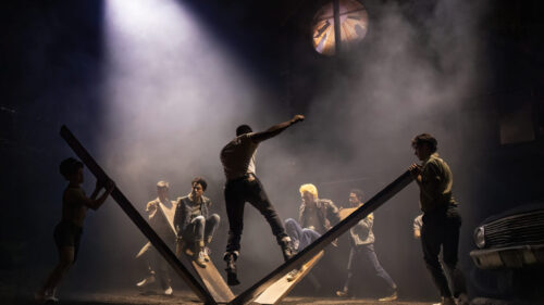 A dramatic scene unfolds with several young people involved in an intense moment. Two men are seated on large wooden planks angled against each other, while another man jumps off the planks with a powerful pose. Other men stand around, watching or holding the planks. The background is dark and smoky, illuminated by a single bright spotlight from above, creating a mysterious and tense atmosphere. A broken circular window at the top adds to the dramatic setting.
