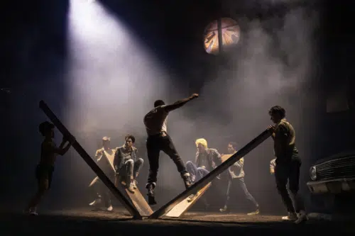 A dramatic scene unfolds with several young people involved in an intense moment. Two men are seated on large wooden planks angled against each other, while another man jumps off the planks with a powerful pose. Other men stand around, watching or holding the planks. The background is dark and smoky, illuminated by a single bright spotlight from above, creating a mysterious and tense atmosphere. A broken circular window at the top adds to the dramatic setting.