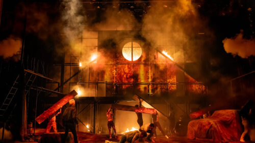 A chaotic and dramatic scene depicts a large fire engulfing a structure, with thick smoke and intense flames lighting up the area. Several young people are scattered around the stage, some carrying large wooden planks, while others are on the ground amidst the debris.