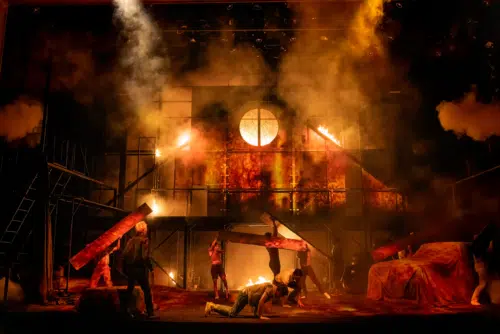A chaotic and dramatic scene depicts a large fire engulfing a structure, with thick smoke and intense flames lighting up the area. Several young people are scattered around the stage, some carrying large wooden planks, while others are on the ground amidst the debris.