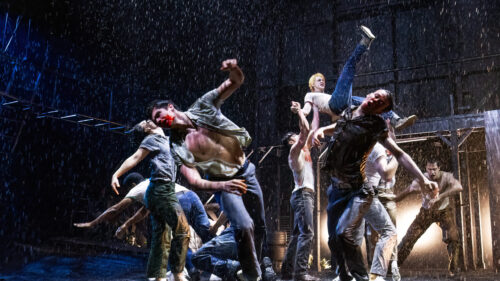 A group of men are engaged in an intense, dramatic fight scene under pouring rain. The men are wearing soaked, casual clothing such as jeans and t-shirts, and some have blood on their faces. One man is being lifted into the air, adding to the chaos and energy of the scene. The background is dark, with rain pouring down, illuminated by stage lights, creating a gritty and action-packed atmosphere.