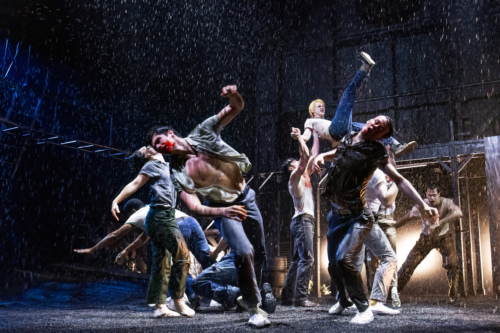 A group of men are engaged in an intense, dramatic fight scene under pouring rain. The men are wearing soaked, casual clothing such as jeans and t-shirts, and some have blood on their faces. One man is being lifted into the air, adding to the chaos and energy of the scene. The background is dark, with rain pouring down, illuminated by stage lights, creating a gritty and action-packed atmosphere.