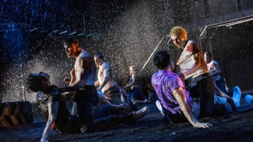 A group of young men are engaged in a violent and chaotic brawl under heavy rain. The scene is intense, with individuals grappling and fighting on the ground, their clothes soaked and stained. Some men have visible injuries and blood on their faces and bodies. The dark, rain-soaked environment is illuminated by dramatic lighting, emphasizing the raw energy and tension of the fight.