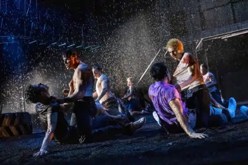 A group of young men are engaged in a violent and chaotic brawl under heavy rain. The scene is intense, with individuals grappling and fighting on the ground, their clothes soaked and stained. Some men have visible injuries and blood on their faces and bodies. The dark, rain-soaked environment is illuminated by dramatic lighting, emphasizing the raw energy and tension of the fight.