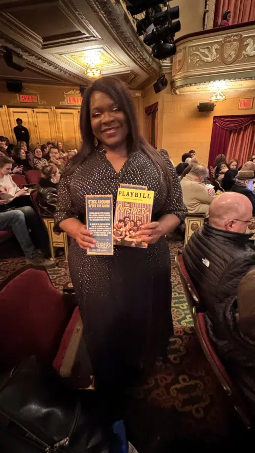 A medium shot of a smiling Black woman with long dark hair, standing in the aisle of the Bernard B. Jacobs Theatre. She is wearing a shimmering, dark-textured dress and holding a "Playbill" and a promotional flyer for the Broadway musical "The Outsiders." The ornate, gold-trimmed interior of the theater and a seated audience are visible in the background.