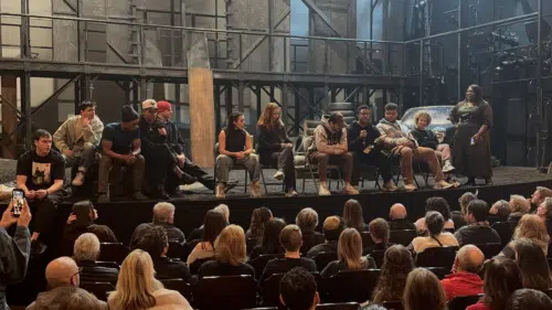 A wide shot from the audience’s perspective showing a "talkback" session on the stage of "The Outsiders." Ten cast members are seated in a row on the gritty, industrial-style set, which features scaffolding, tires, and a rusted car. A moderator stands to the far right, holding a microphone. The foreground shows the backs of the heads of a nearly full audience in the darkened theater.