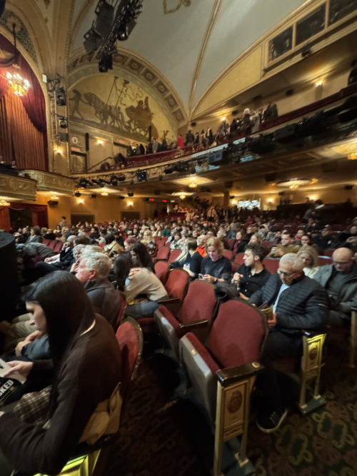 A wide-angle view of the interior of the Bernard B. Jacobs Theatre from a side aisle. The image captures the red velvet seating filled with audience members, the glowing chandeliers, and the ornate architectural details of the balcony. A large, classical-style mural is visible on the arched wall above the box seats.